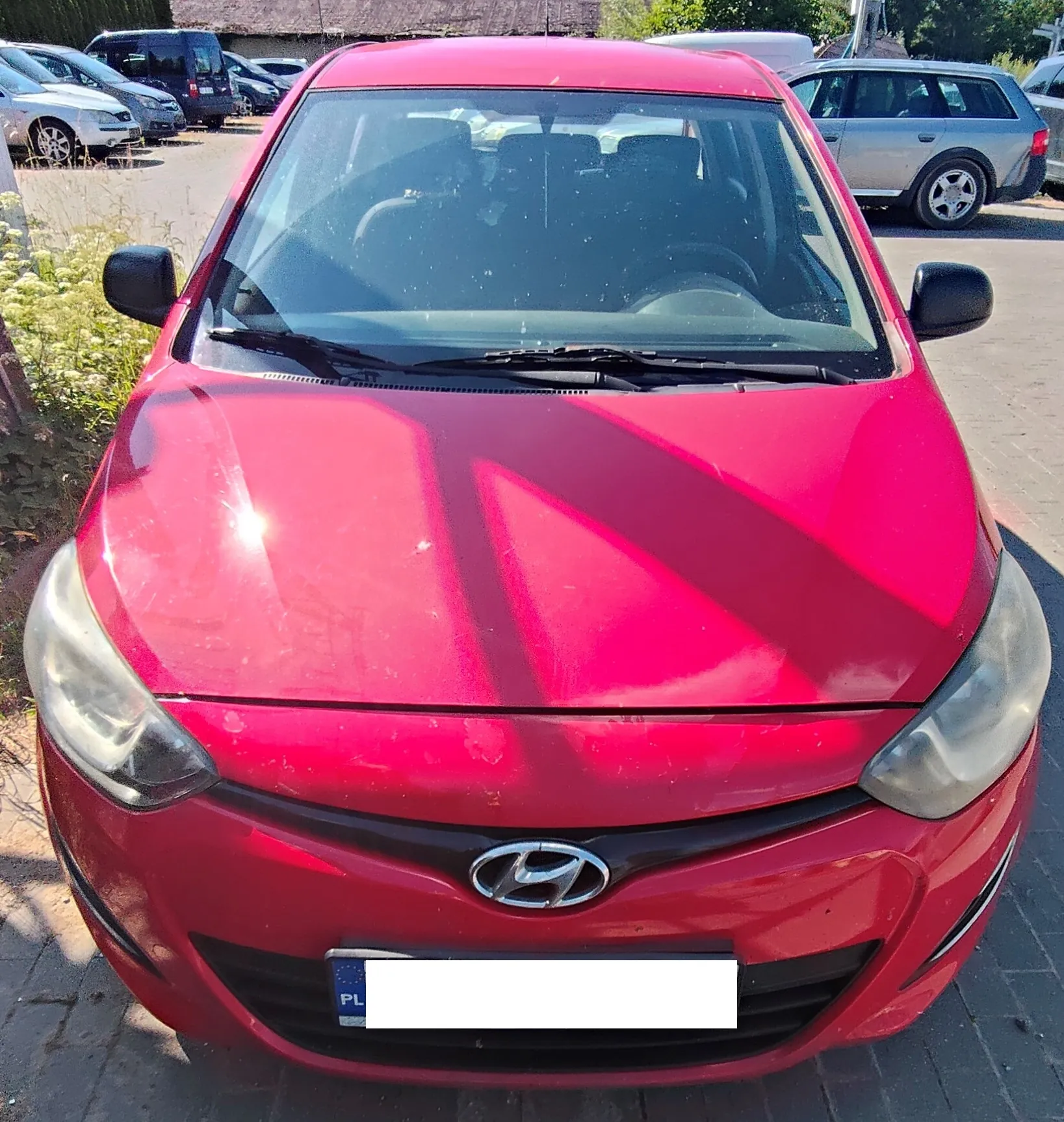 Widok z przodu czerwonego samochodu marki Hyundai zaparkowanego na placu. Auto ma widoczne logo producenta na masce oraz polską tablicę rejestracyjną (zasłoniętą). W tle widoczne są inne zaparkowane samochody, w tym srebrny kombi po prawej stronie oraz ciemne vany po lewej.