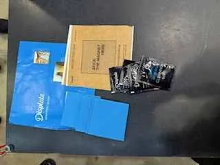 Na ciemnym stole leżą różne przedmioty: niebieska koperta z napisem "Diplom", brązowa koperta lub folder z częściowo widocznym tekstem, niebieskie arkusze papieru oraz stos czarno-białych zdjęć lub małych broszur. W lewym dolnym rogu widoczny jest fragment stopy osoby.