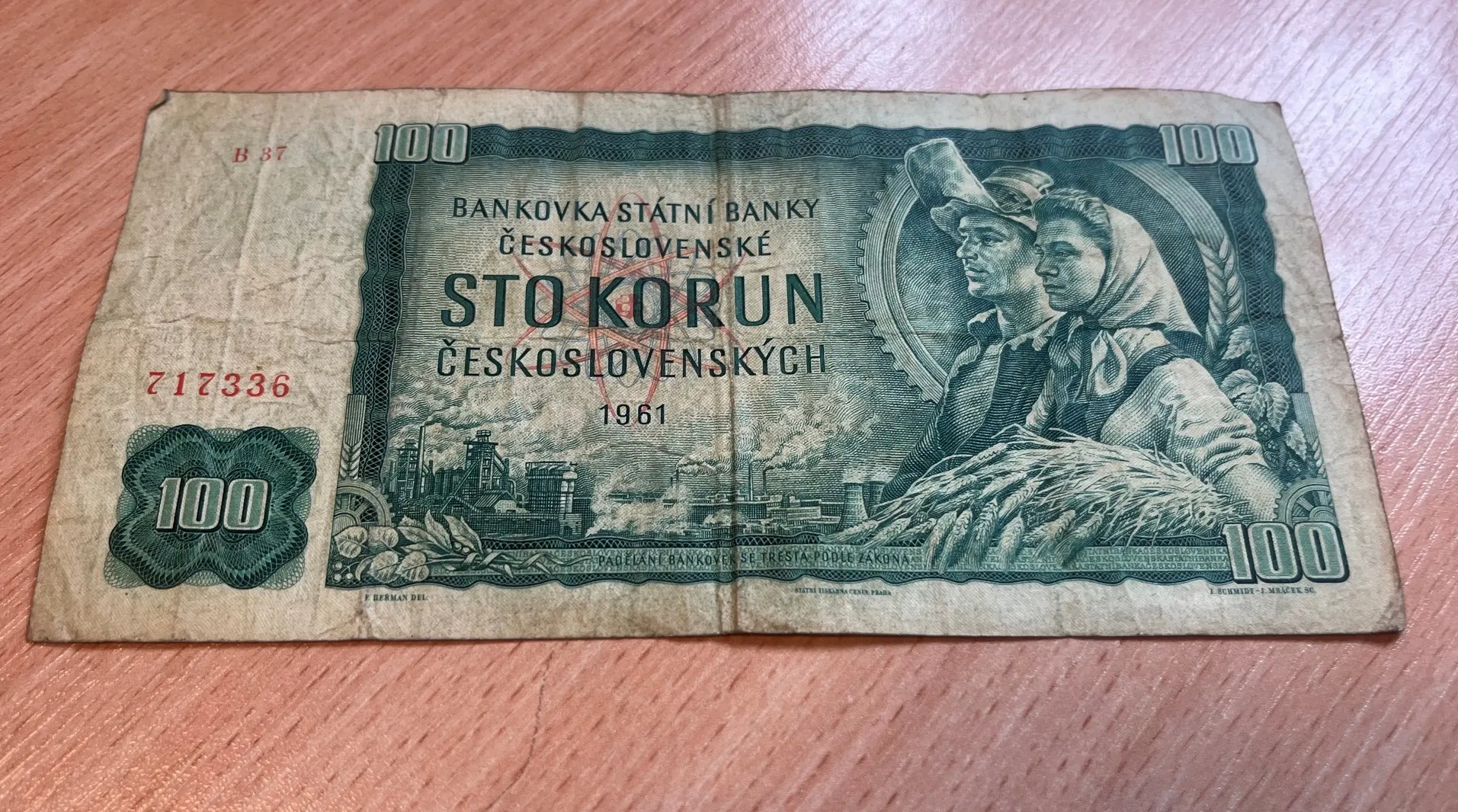 Stary, pognieciony banknot o nominale 100 koron czechosłowackich z 1961 roku. Banknot jest w kolorze zielonym i beżowym. W centralnej części znajduje się napis „BANKOVKA STÁTNÍ BANKY ČESKOSLOVENSKÉ STO KORUN ČESKOSLOVENSKÝCH 1961”. Po prawej stronie widnieje portret dwojga ludzi: mężczyzny w kapeluszu i kobiety w chustce na głowie. Po lewej stronie znajduje się grafika przedstawiająca zakład przemysłowy z kominami. W rogach widnieje liczba 100, a po lewej stronie czerwony numer seryjny 717336.