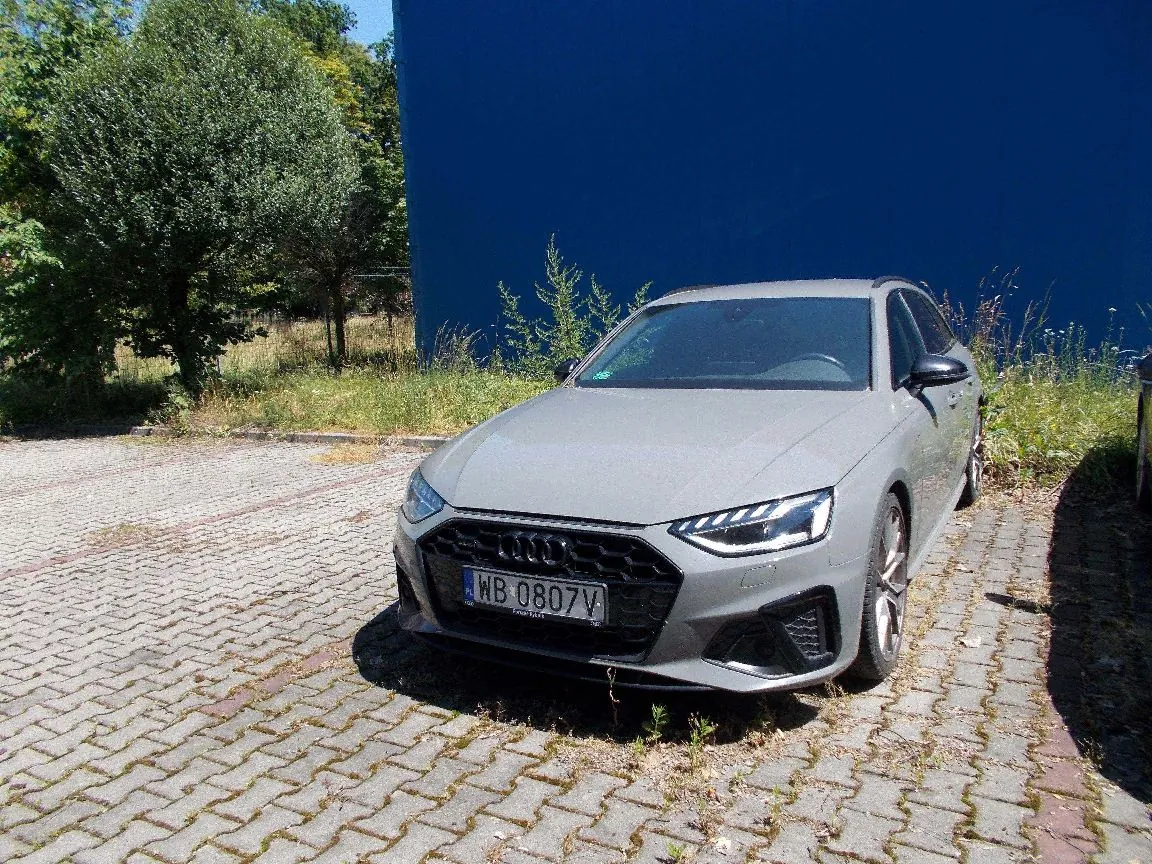 Srebrnoszary samochód marki Audi zaparkowany na kostce brukowej. Pojazd ma polską tablicę rejestracyjną o numerze WB 0807V. W tle znajduje się wysoka, niebieska ściana oraz zielone drzewa i krzewy.