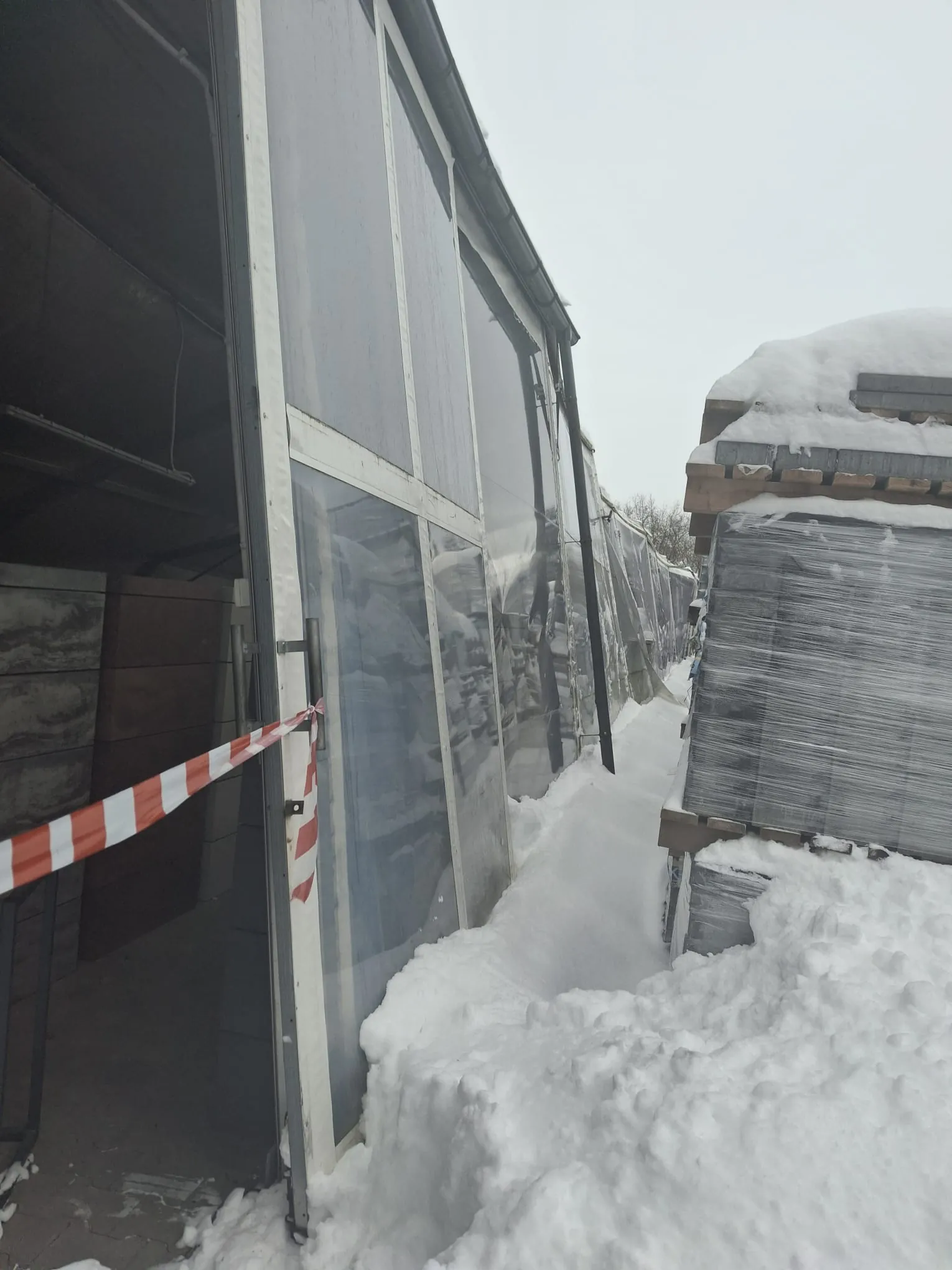 Zdjęcie przedstawia zimowy krajobraz z budynkiem o szklanych ścianach i białej ramie po lewej stronie. Przed wejściem do budynku znajduje się czerwono-biała taśma ostrzegawcza. Po prawej stronie widoczne są stosy materiałów owiniętych folią, pokryte śniegiem. Cały teren jest pokryty grubą warstwą śniegu, a na tle szare, zachmurzone niebo.