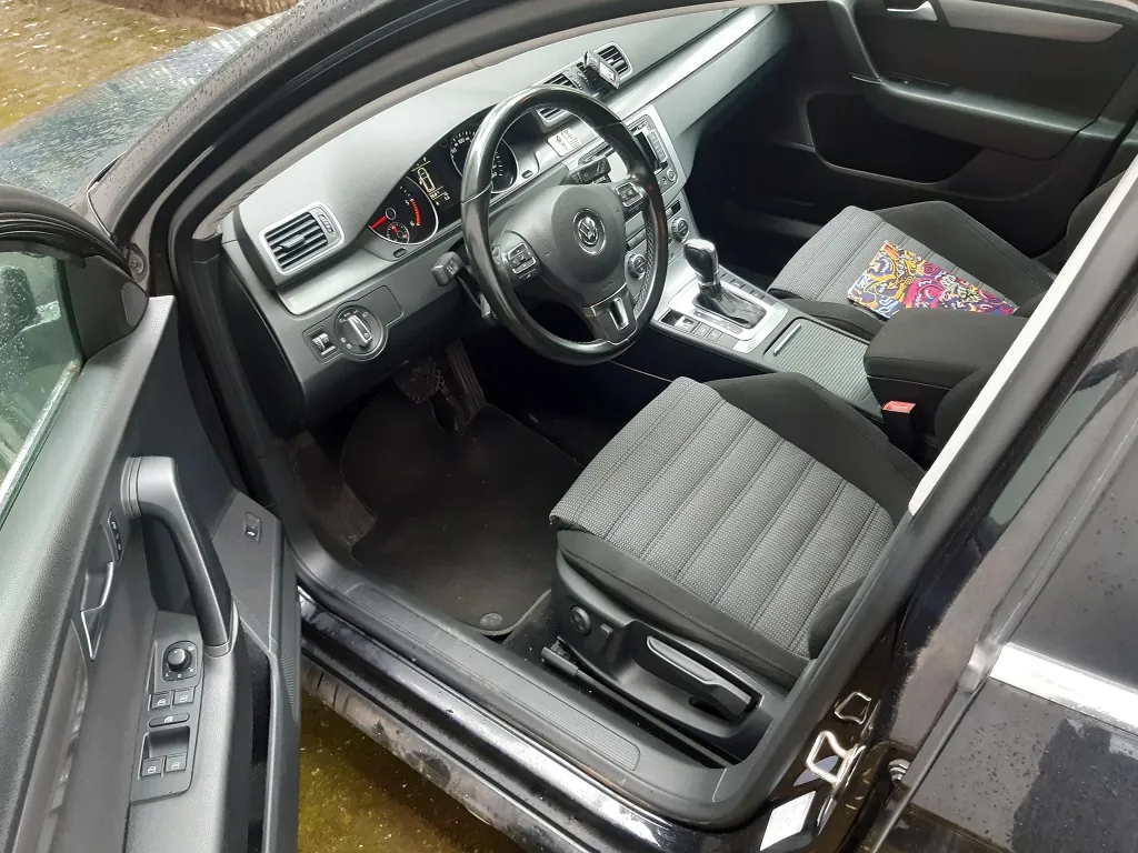 Wnętrze samochodu widziane z otwartych drzwi kierowcy. Widoczna czarna tapicerka, kierownica z logo Volkswagena, deska rozdzielcza z zegarami, konsola środkowa z dźwignią zmiany biegów oraz dwa przednie fotele z szarymi wstawkami. Na siedzeniu pasażera leży kolorowa, wzorzysta tkanina.