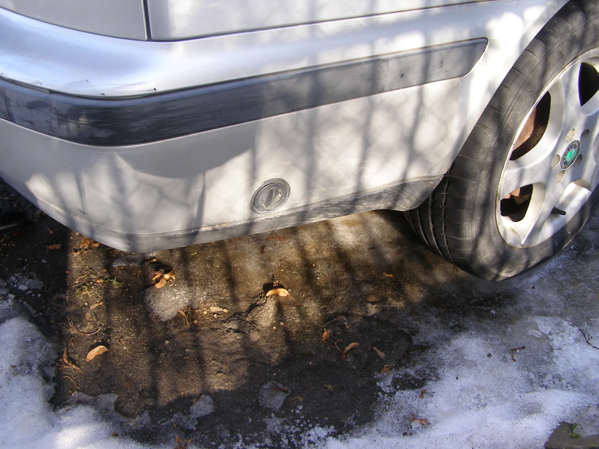 Zbliżenie przedniej części srebrnego samochodu, obejmujące zderzak, przednie koło z felgą i oponą. Na zderzaku widoczny okrągły czujnik. Podwozie i koło znajdują się nad nawierzchnią z resztkami śniegu, lodu oraz rozsypanymi suchymi liśćmi. Na felgach widoczny zielony emblemat. Cień siatki lub ogrodzenia rzuca pionowe i poziome linie na samochód i podłogę.