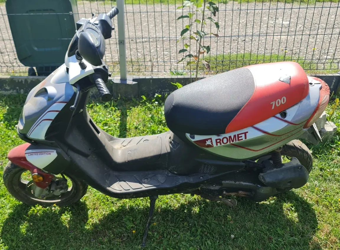 Skuter marki Romet w kolorach srebrnym, czerwonym i czarnym, zaparkowany na trawie przed metalowym ogrodzeniem. Na tylnej części skutera widnieje napis 'ROMET' oraz liczba '700'. Na przedniej owiewce znajduje się napis 'LIMITED EDITION'. Pojazd stoi na bocznej stopce.