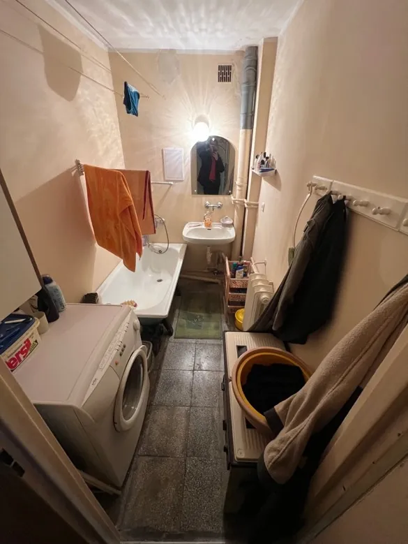 Wnętrze łazienki z białą wanną, umywalką z lustrem, pralką, pomarańczowym i niebieskim ręcznikiem na suszarce, akwarium, wiszącymi ubraniami na wieszakach, płytkami na ścianach oraz różnymi przedmiotami na półkach. Oświetlenie z żarówką na ścianie.