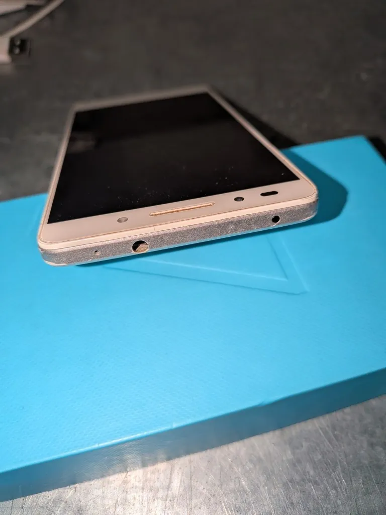 Biały smartfon z czarnym ekranem leżący na niebieskim, prostokątnym pudełku. Urządzenie jest pokazane pod kątem, widoczna jest jego górna krawędź z gniazdem słuchawkowym oraz przedni panel z głośnikiem i czujnikami.