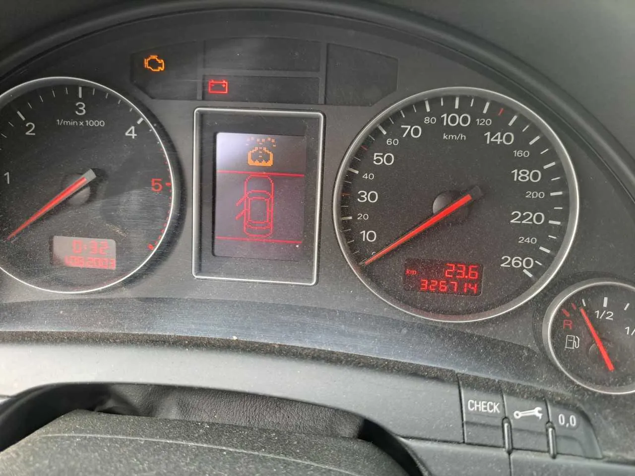 Zdjęcie przedstawia deski rozdzielcze samochodu. Po lewej stronie znajduje się tachometr z czerwoną strzałką wskazującą około 1,5 (1/min x 1000), poniżej cyfrowy wyświetlacz z czerwonymi cyframi 0:32 i 1062003. W centrum ekran z czerwonym symbolem samochodu i ostrzeżeniem w postaci ikony. Po prawej stronie prędkościomierz z zakresem do 260 km/h, strzałka wskazuje 0, poniżej cyfrowy wyświetlacz z wartościami 23,6 i 326714. W prawym dolnym rogu wskaźnik paliwa z czerwoną strzałką na poziomie około 1/2. W górnej części deski widoczne są pomarańczowe i czerwone lampki ostrzegawcze (m.in. symbol silnika i baterii). Na dole panelu przyciski z napisem CHECK oraz ikoną klucza. Całość w kolorze czarnym z czerwonymi i białymi oznaczeniami.