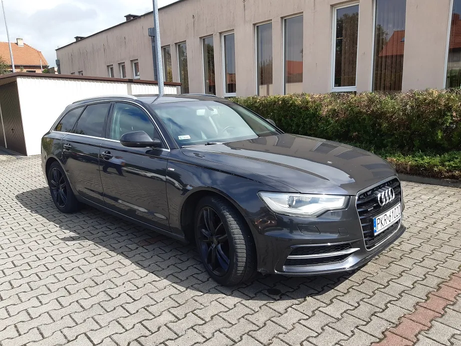 Czarny samochód marki Audi w nadwoziu kombi, zaparkowany na kostce brukowej przed jasnym, nowoczesnym budynkiem z dużymi oknami. Samochód ma czarne felgi i tablicę rejestracyjną o numerze PKR 6102D. W tle widoczny jest niski żywopłot oraz fragment dachu innego budynku.