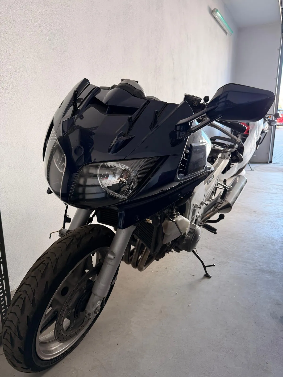 Zbliżenie na przód ciemnogranatowego motocykla sportowego zaparkowanego w jasnym garażu. Widoczna jest przednia opona, reflektor oraz owiewka. Motocykl stoi na stopce bocznej.