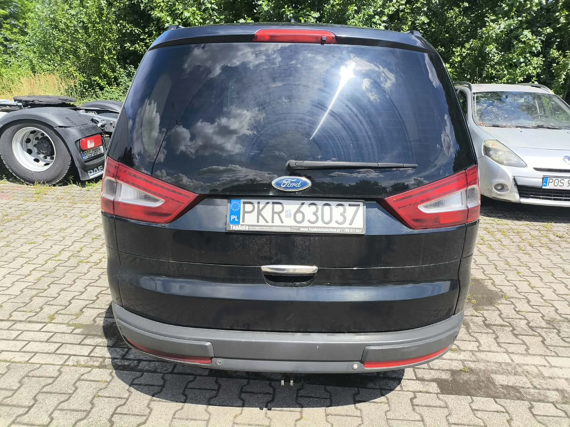 Tylny widok czarnego samochodu marki Ford. Na samochodzie znajduje się polska tablica rejestracyjna o numerze PKR 63037. Poniżej tablicy widnieje napis 'TopAuto' oraz adres strony internetowej i numer telefonu. Samochód zaparkowany jest na kostce brukowej, w tle widoczne są zielone krzewy oraz fragmenty innych pojazdów: ciężarówki po lewej i srebrnego vana po prawej.