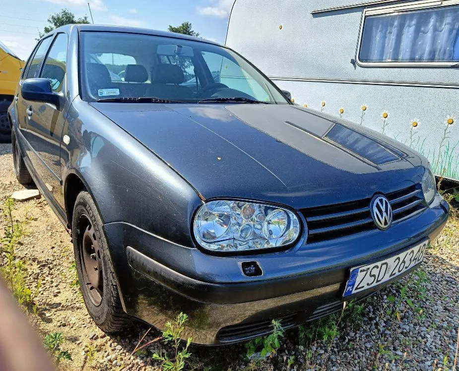 Czarny samochód osobowy marki Volkswagen Golf zaparkowany na żwirowym podłożu. Widoczny jest przód i lewy bok auta. Samochód ma polską tablicę rejestracyjną o numerze ZSD 2634D. W tle znajduje się jasna przyczepa kempingowa z dekoracją w postaci białych kwiatów na boku.