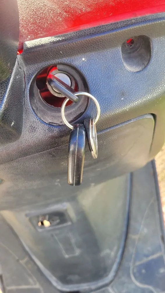 Zbliżenie na czarną, plastikową obudowę skutera z włożonym do stacyjki kluczykiem. Do kluczyka przymocowany jest drugi klucz oraz czarny brelok. Poniżej znajduje się schowek lub klapka z dodatkowym zamkiem.