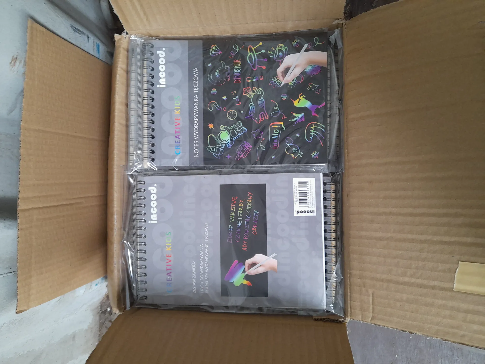 Otwarte kartonowe pudełko zawierające dwa spiralowane notatniki w przezroczystej folii. Górnym notatnikiem jest czarna okładka z kolorowymi holograficznymi rysunkami (dinozaur, jednorożec, astronauta, kwiaty, serca), napisem "DINOSAUR" oraz widoczną ręką trzymającą stylus. Na okładce widnieje logo "incood.", napisy "CREATIVE KIDS" i "NOTES WYDRAPYWANKA TĘCZOWA". Dolny notatnik ma szarą okładkę z czarnym prostokątem zawierającym tekst w języku polskim: "ZDRAP WARSTWĘ CZARNEJ FARBY, ABY POWSTAŁ CIEKAWY OBRAZEK", obok ręki z kolorowym rysikiem. Widoczne są również logo "incood.", napis "CREATIVE KIDS" oraz informacja: "ZESTAW ZAWIERA: 1 RYSIK DO WYDRAPYWANIA, 10 ARKUSZY WYDRAPYWANEK TĘCZOWYCH". Obok znajduje się kod kreskowy.