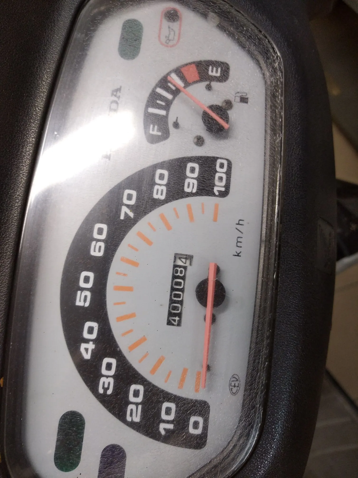 Zbliżenie na analogowy licznik prędkości i wskaźnik poziomu paliwa w pojeździe. Prędkościomierz ma skalę od 0 do 100 km/h, a aktualny przebieg na liczniku kilometrów wynosi 400084. Powyżej znajduje się mniejszy wskaźnik paliwa z oznaczeniami F (pełny) i E (pusty). Całość jest osłonięta przez porysowaną szybkę w czarnej obudowie.