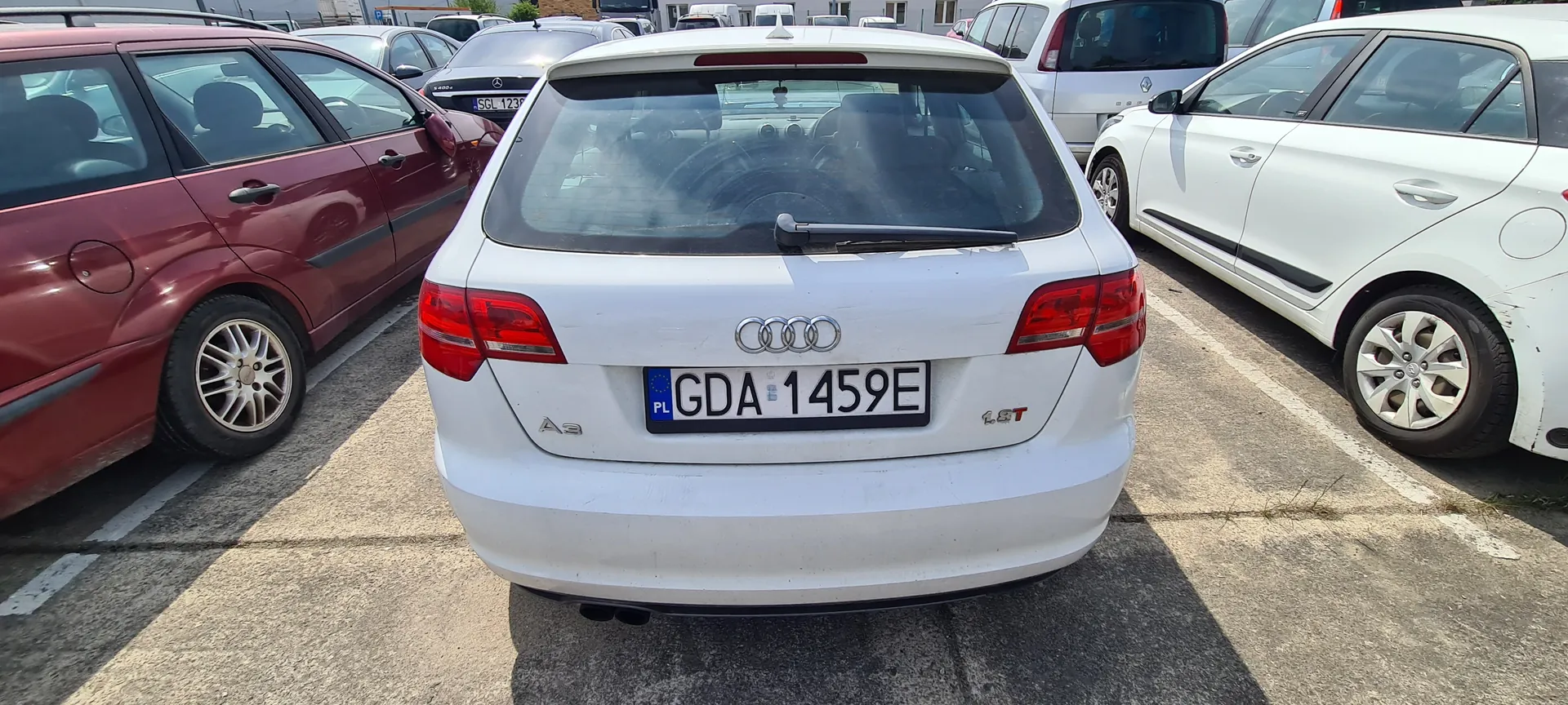 Biały samochód Audi A3 z tablicą rejestracyjną GDA 1459E (kod kraju PL) zaparkowany na betonowym parkingu. Widoczne są cztery pierścienie logo Audi, oznaczenie modelu A3 oraz 1.8T na tylnej części pojazdu. Po obu stronach stoją inne samochody: czerwony po lewej i biały po prawej. Tło zawiera dodatkowe zaparkowane pojazdy oraz budynki.