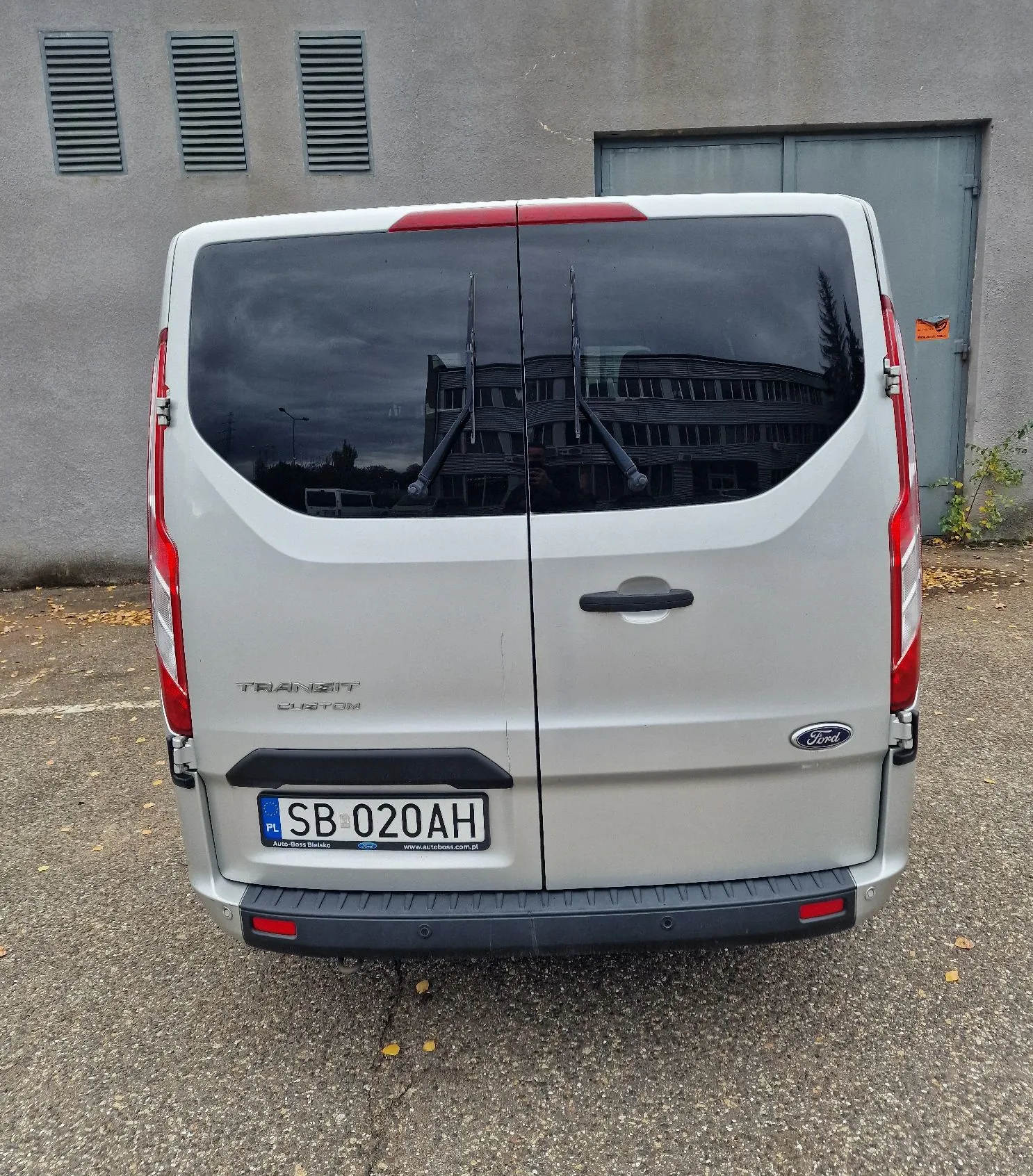 Tylny widok białego vana marki Ford Transit Custom zaparkowanego na asfaltowej nawierzchni przed szarym budynkiem. Pojazd posiada dwie tylne szyby z wycieraczkami, polską tablicę rejestracyjną o numerze SB 020AH oraz logo Forda. Na klapie bagażnika widnieje napis TRANSIT CUSTOM.