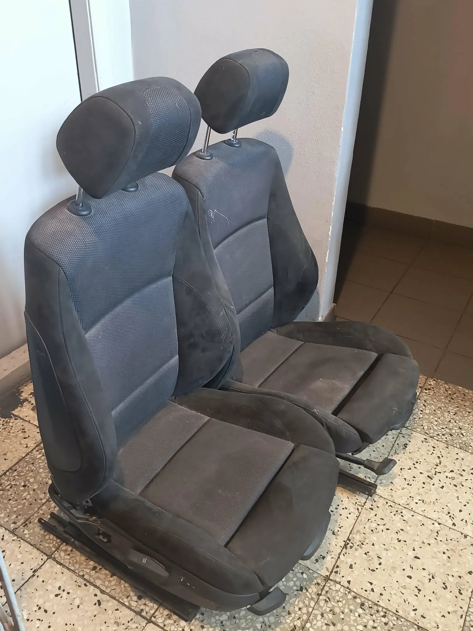 Dwa czarne, materiałowe fotele samochodowe ustawione obok siebie na jasnej, kropkowanej podłodze w pomieszczeniu. Fotele posiadają regulowane zagłówki i widoczne są na nich ślady użytkowania oraz kurz.