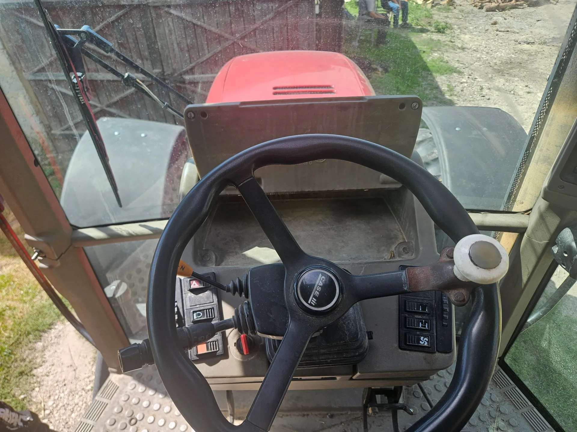 Widok z wnętrza kabiny ciągnika rolniczego. Na pierwszym planie znajduje się czarna kierownica z logo 'CASE IH' na środku. Za kierownicą widoczna jest deska rozdzielcza z przełącznikami i przyciskami. Przez przednią szybę widać czerwoną maskę ciągnika oraz fragmenty otoczenia na zewnątrz.