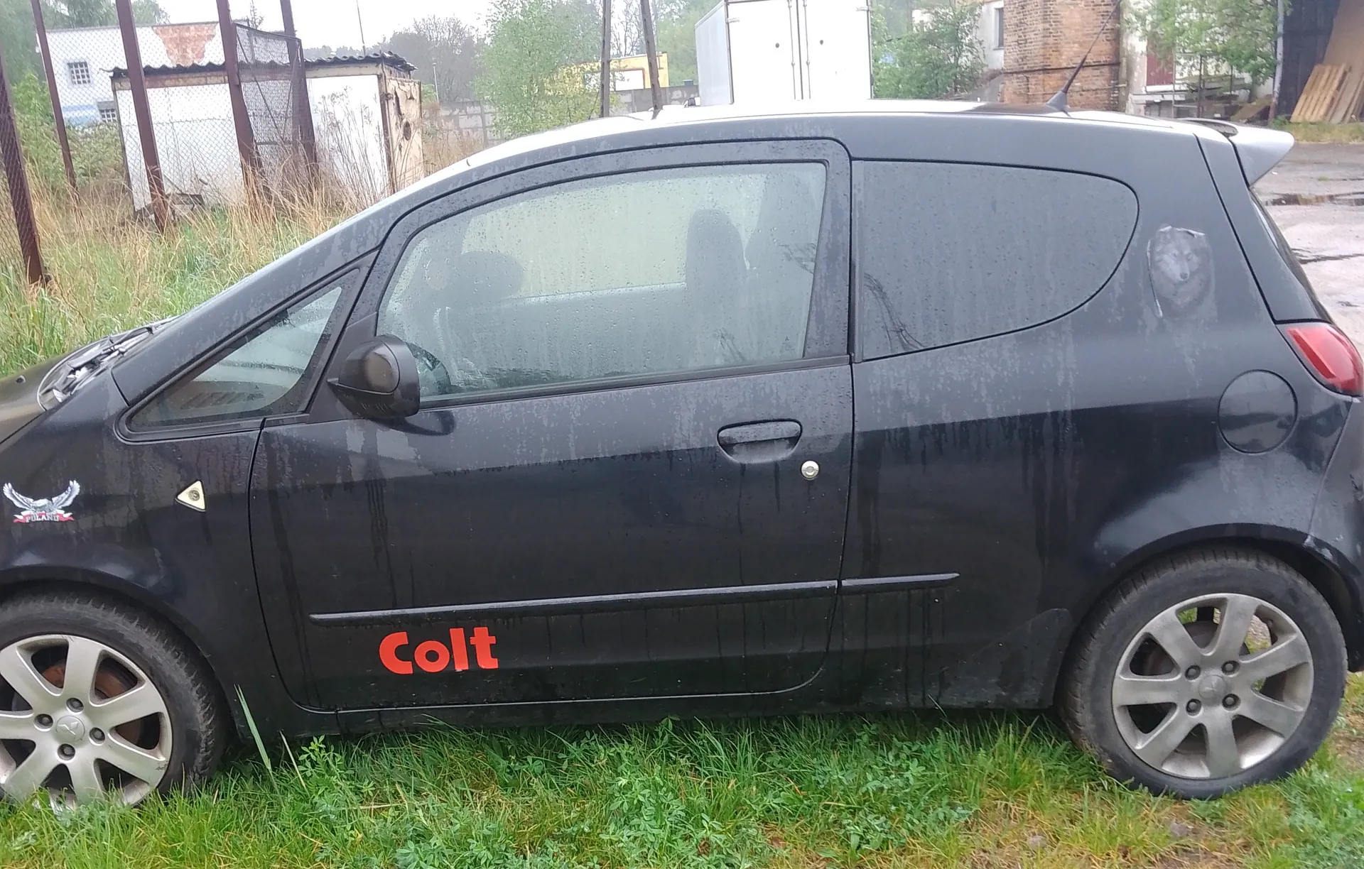 Czarny samochód osobowy Mitsubishi Colt z czerwonym napisem "Colt" na boku, zaparkowany na trawie. Na przednim błotniku widoczny jest naklejony emblemat z orłem. Tło stanowią budynki, ogrodzenie i wysokie trawy. Samochód pokryty jest kroplami wody, a na tylnym szybie widoczny jest naklejony obrazek zwierzęcia. Koła mają srebrne felgi.