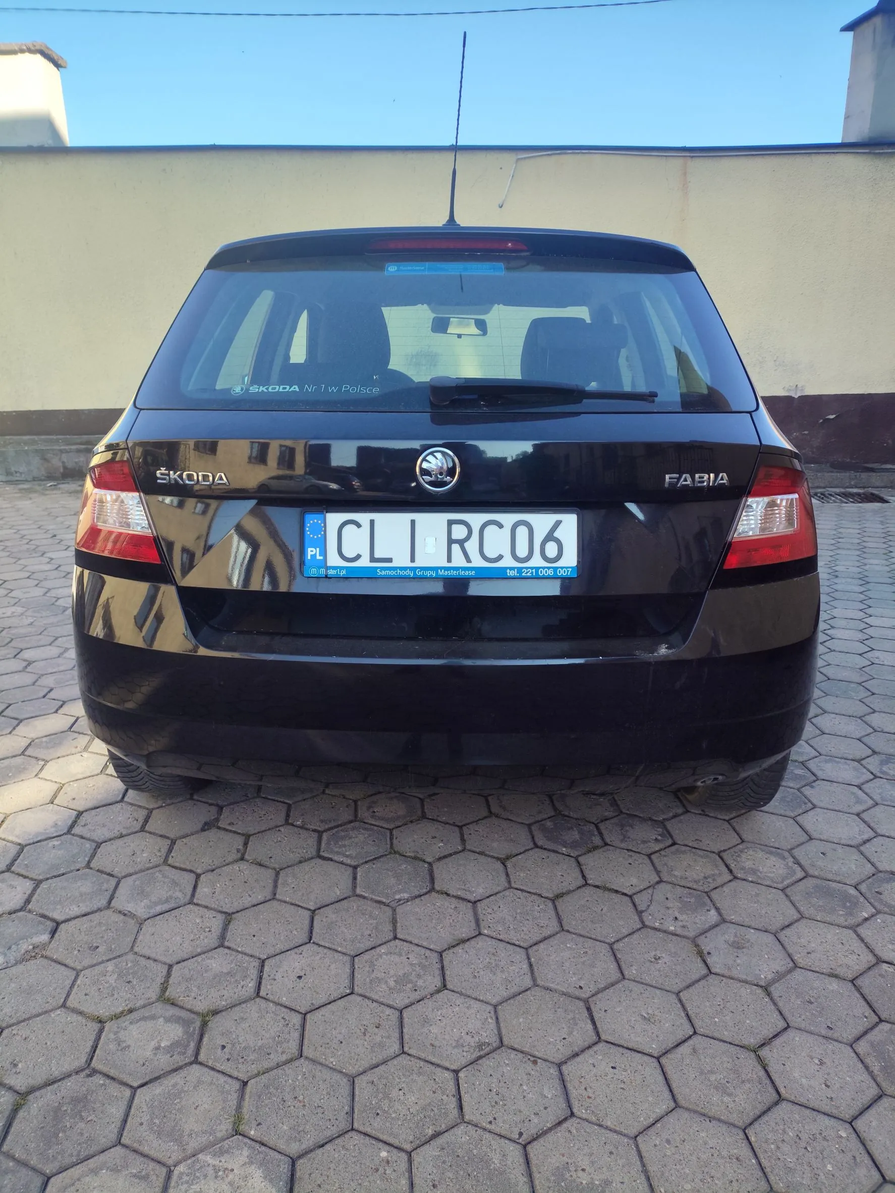 Tylny widok czarnego samochodu marki Skoda Fabia zaparkowanego na kostce brukowej o heksagonalnym kształcie. Samochód posiada polską tablicę rejestracyjną o numerze CLI RC06. Na klapie bagażnika widoczne są napisy „ŠKODA” po lewej stronie oraz „FABIA” po prawej, a w centrum logo marki. Na tylnej szybie znajduje się naklejka z tekstem „ŠKODA Nr 1 w Polsce”. W tle widoczny jest jasny mur pod błękitnym niebem.