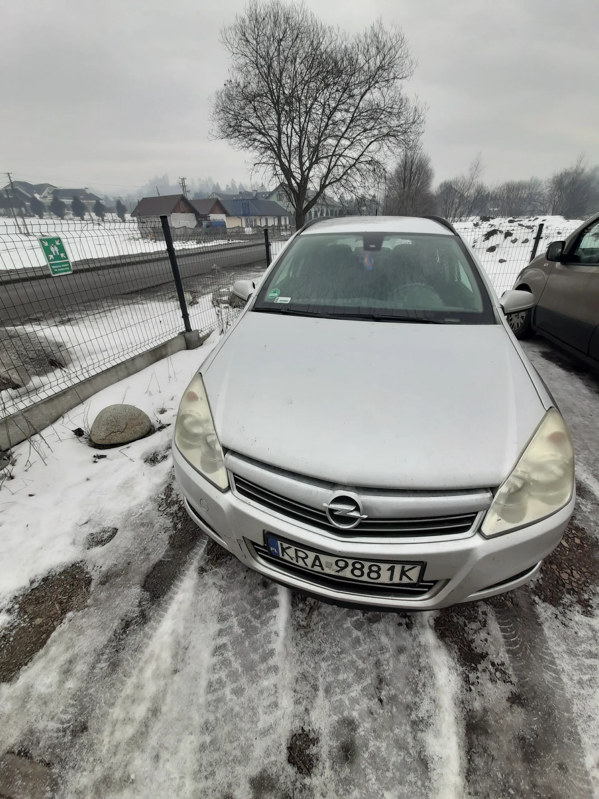Srebrny samochód z tablicą rejestracyjną KRA 9881K zaparkowany na śnieżnej powierzchni z widocznymi plamami ziemi. W tle częściowo widoczny szary samochód, metalowe ogrodzenie, drzewa bez liści, zielony znak drogowy oraz budynki. Niebo jest zachmurzone.