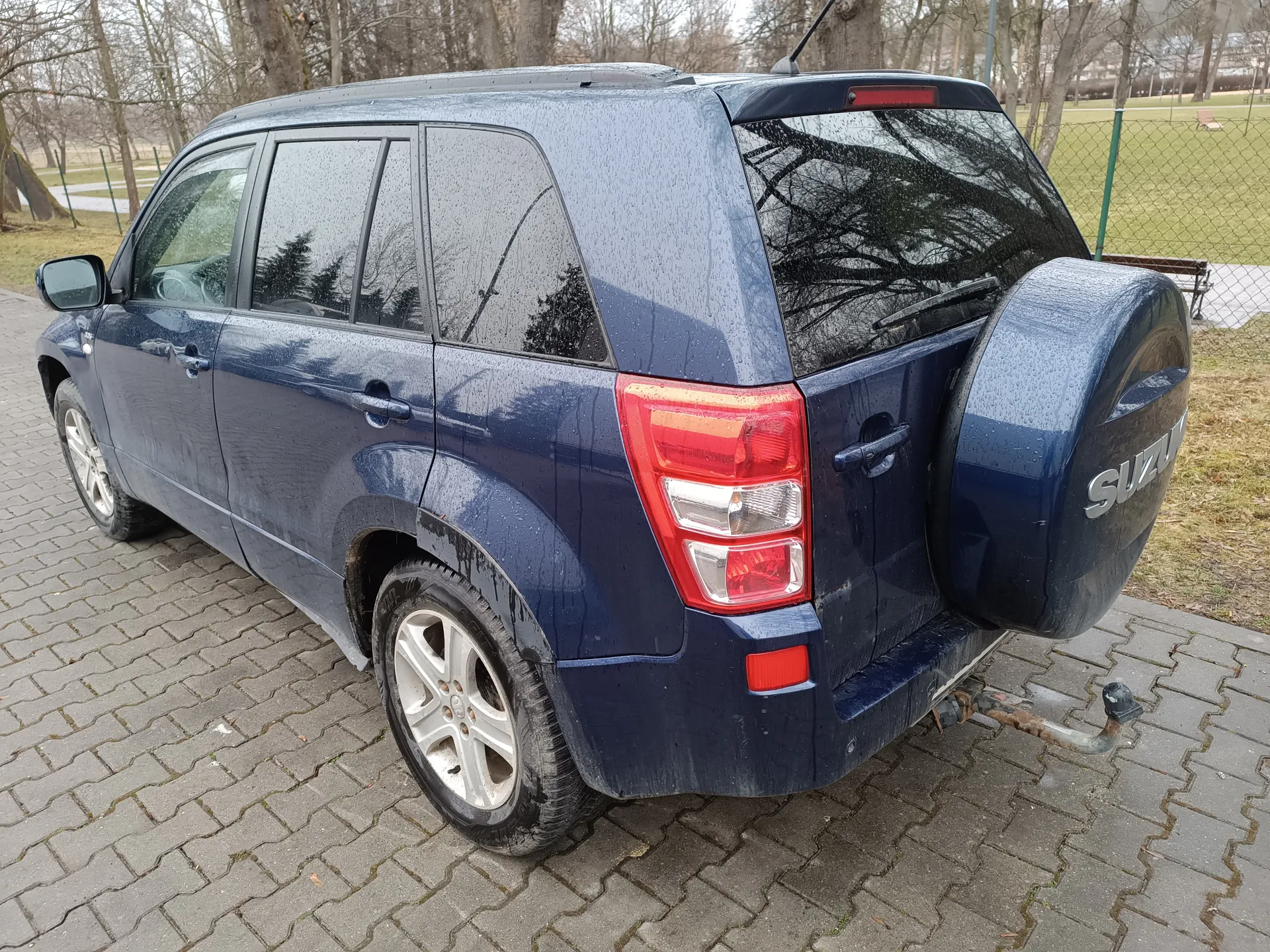 Ciemnoniebieski SUV marki Suzuki z widocznym na zapasowym kole napisem 'SUZUKI', zaparkowany na brukowanej powierzchni. Samochód pokryty jest kroplami deszczu, widoczne są czerwone światła tylne, srebrne felgi oraz hak holowniczy. W tle: ogrodzenie siatkowe, ławka, drzewa bez liści i trawnik.