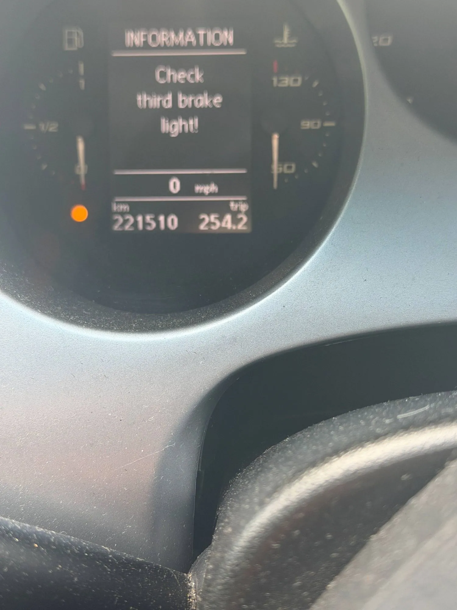 Zbliżenie na wyświetlacz komputera pokładowego w samochodzie. Na ekranie widnieje napis 'INFORMATION' oraz komunikat ostrzegawczy: 'Check third brake light!'. Poniżej znajduje się licznik prędkości wskazujący '0 mph', przebieg pojazdu '221510 km' oraz licznik podróży 'trip 254.2'. Po lewej stronie świeci się pomarańczowa kontrolka.