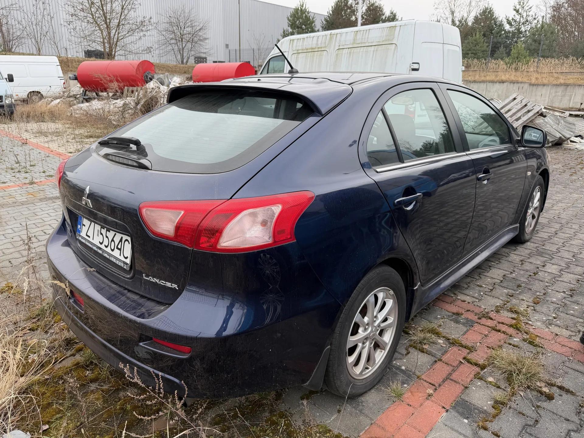 Ciemnoniebieski samochód Mitsubishi Lancer zaparkowany na brukowanej powierzchni. Widok z tyłu, z widoczną tablicą rejestracyjną PL ZI 5564C oraz napisem "LANCER" na bagażniku. W tle białe furgony, czerwone zbiorniki, betonowa ściana, suche trawy i drzewa bez liści. Na ziemi widoczne są plamy mchu między kostkami brukowymi.