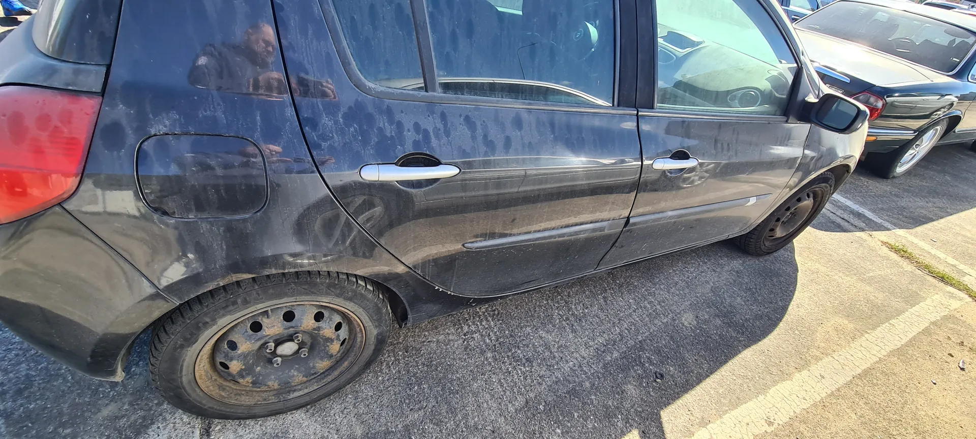 Czarny samochód osobowy zaparkowany na asfaltowej powierzchni. Widoczne są brudne boczne drzwi z srebrnymi uchwytami, zatłuszczony tylny reflektor, zanieczyszczona karoseria z plamami oraz koła z rdzawymi felgami. W tle drugi samochód i białe linie parkingu. Na szybie odbicie osoby fotografującej.