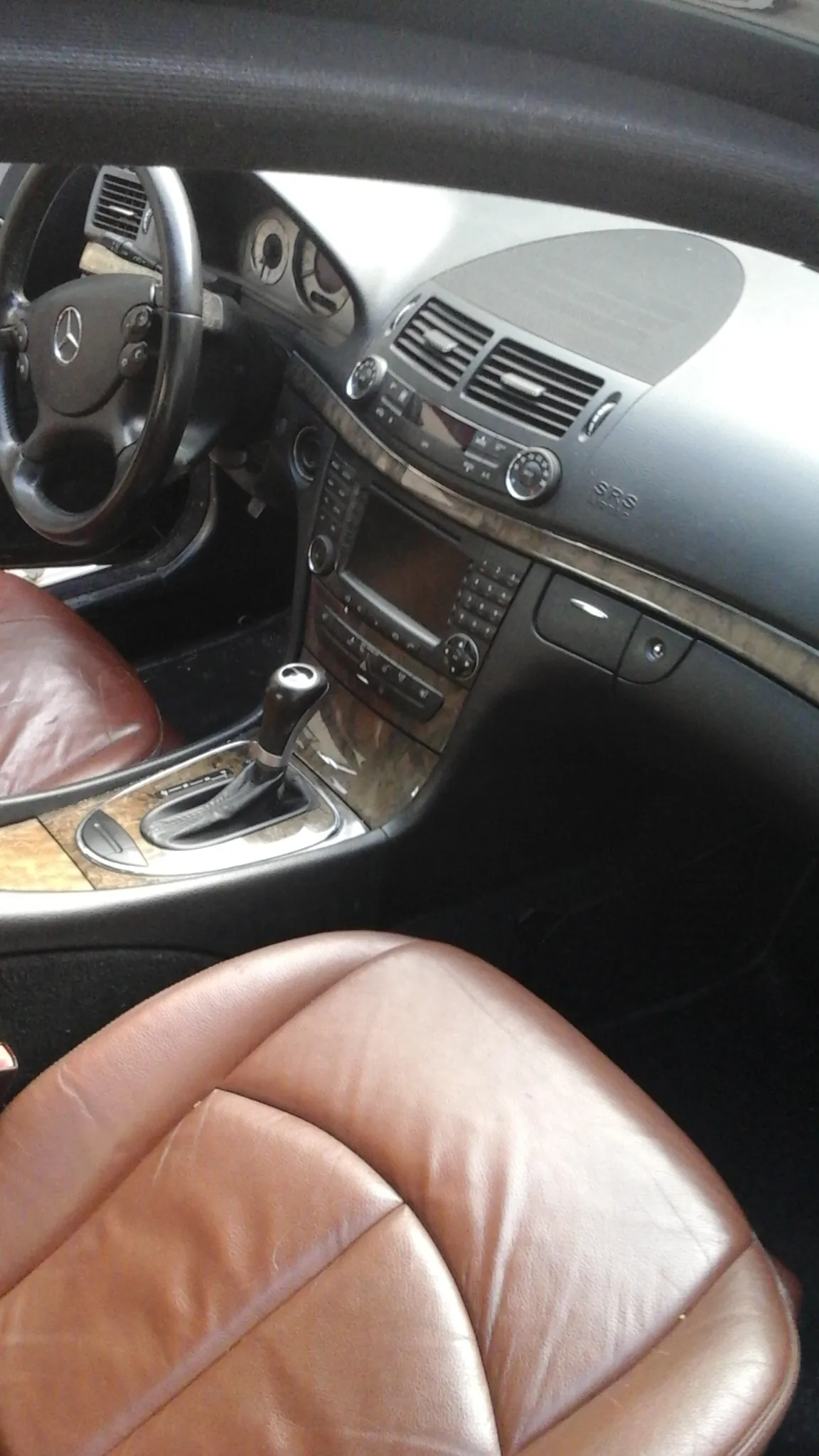 Wnętrze samochodu marki Mercedes z widoczną kierownicą z logo, deską rozdzielczą z wentylacjami, przyciskami, ekranem, dźwignią zmiany biegów oraz brązowym skórzonym siedzeniem. Deska rozdzielcza posiada wykończenie z drewna.