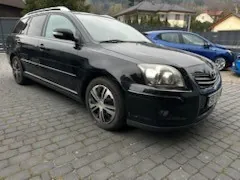 Czarny samochód typu kombi zaparkowany na brukowanej powierzchni. W tle widoczny niebieski samochód oraz domy z dachami. Samochody są ustawione prosto, horyzont jest poziomy.