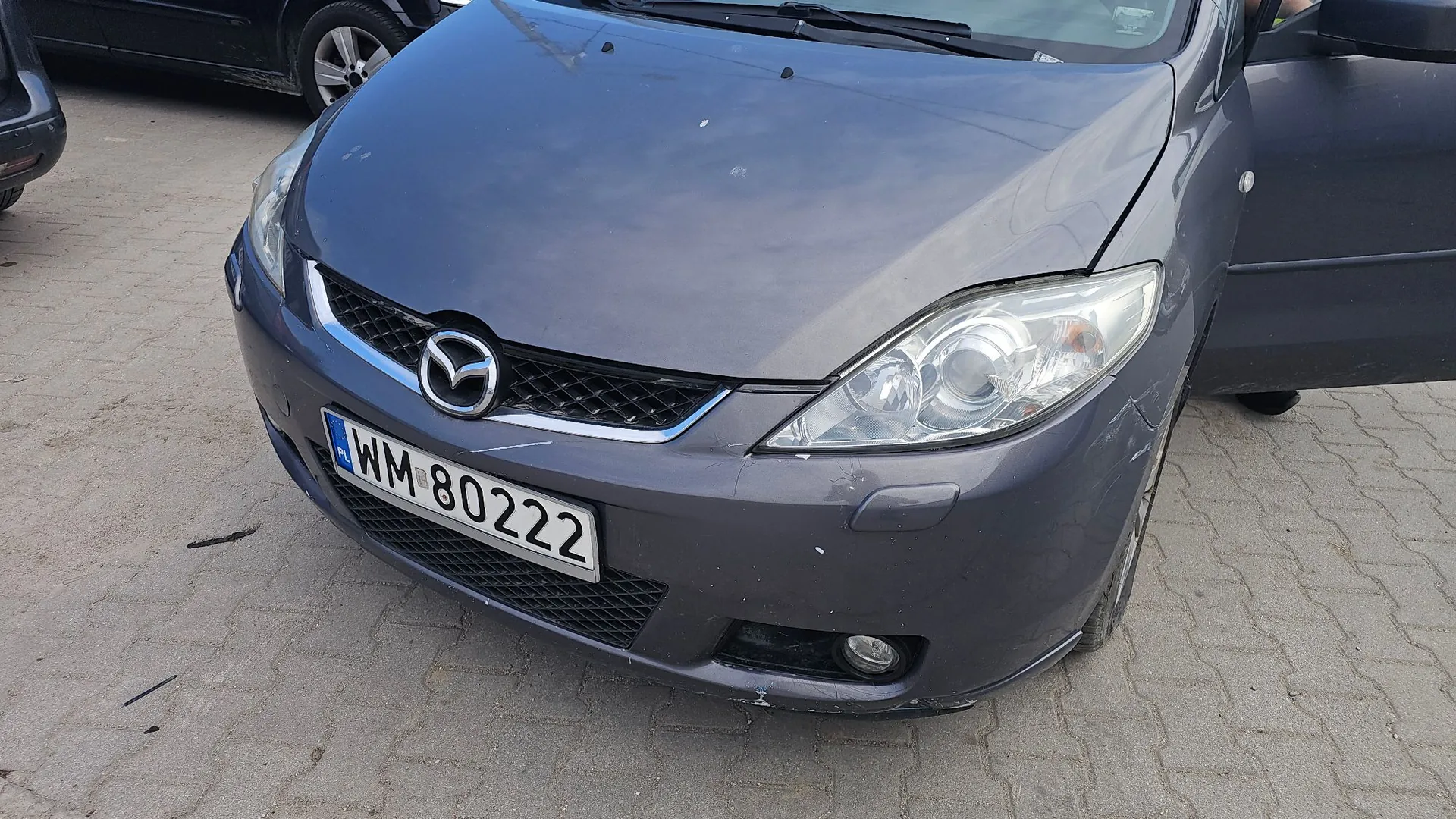 Przednia część szarego samochodu marki Mazda. Widoczny jest grill z logo producenta, reflektory oraz polska tablica rejestracyjna o numerze WM 80222. Na prawym błotniku samochodu widoczne są zarysowania. Pojazd stoi na kostce brukowej.