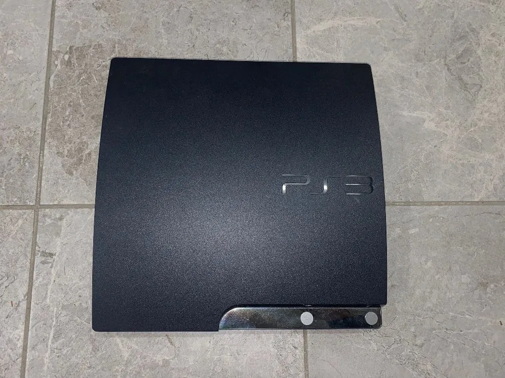 Czarna konsola PlayStation 3 leżąca na szarych, kamiennych płytkach podłogowych. Na obudowie widnieje wytłoczony napis 'PS3'. W dolnej części urządzenia znajduje się srebrny, błyszczący pasek z dwoma okrągłymi otworami.