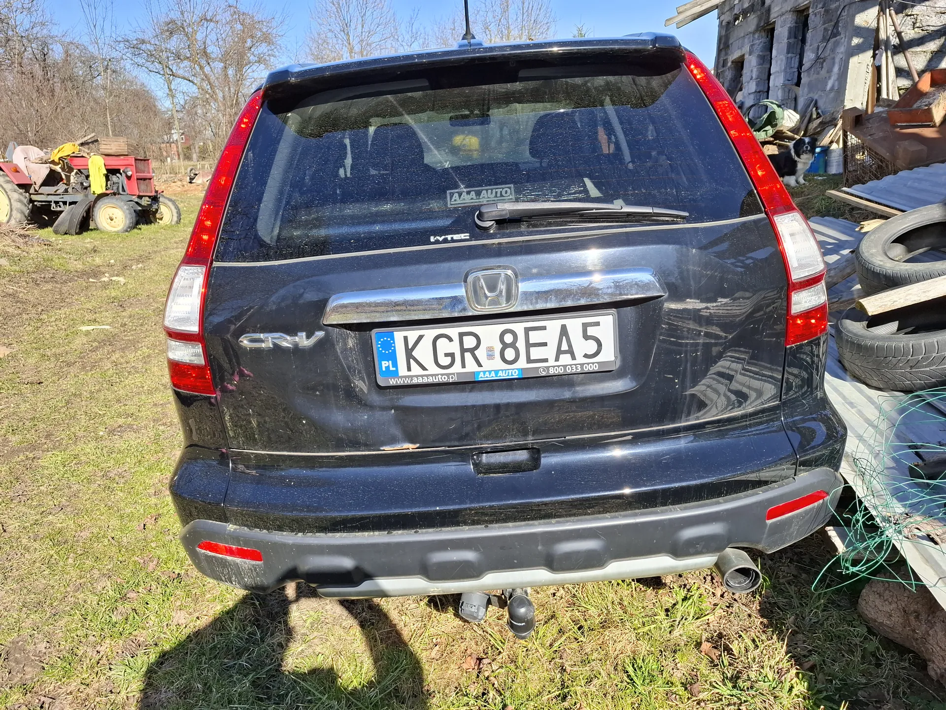 Tył czarnego samochodu Honda CR-V z tablicą rejestracyjną KGR 8EA5. Na tablicy widoczne są elementy: PL, www.aaaauto.pl, AAA AUTO oraz numer 800 033 000. Na szybie tylnej naklejka z napisem AAA AUTO. W tle trawa, czerwony ciągnik, stos opon, nieukończony budynek z betonowych bloków, pies oraz stosy materiałów budowlanych. Niebo jest bezchmurne.