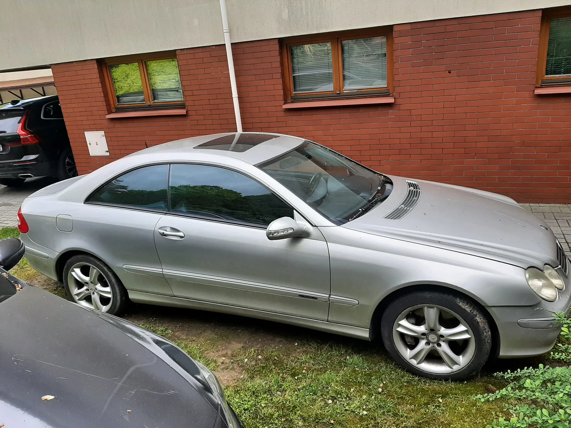 Srebrny samochód osobowy typu coupé marki Mercedes-Benz zaparkowany na trawie przed budynkiem z czerwonej cegły. Budynek posiada okna z drewnianymi ramami i białą rynną. W tle widoczny jest czarny samochód zaparkowany pod daszkiem. Na pierwszym planie fragment czarnego pojazdu. Samochód srebrny ma czarne felgi, przednie reflektory i dach z szybą panoramiczną. Trawa wokół pojazdu jest zielona z widocznymi plamami ziemi.