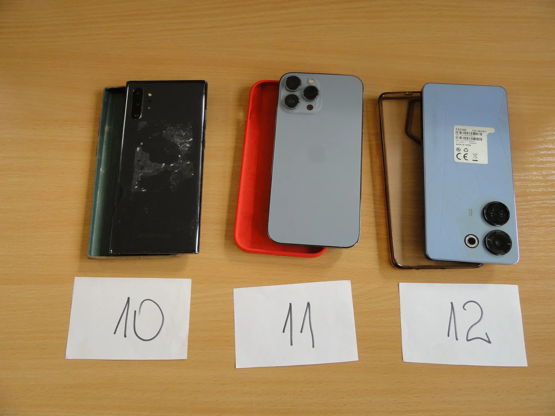 Trzy smartfony ułożone na drewnianym stole. Po lewej czarny smartfon z uszkodzonym tyłem w zielonym etui, pod nim kartka z napisem 10. W środku szary smartfon z logo Apple i trzema aparatami na czerwonym etui, pod nim kartka z napisem 11. Po prawej jasnoniebieski smartfon z naklejką zawierającą kod kreskowy i symbole CE, z dwoma aparatami w przezroczystym etui, pod nim kartka z napisem 12. Wszystkie smartfony są ułożone pionowo, a kartki z numerami umieszczone są pod nimi poziomo.