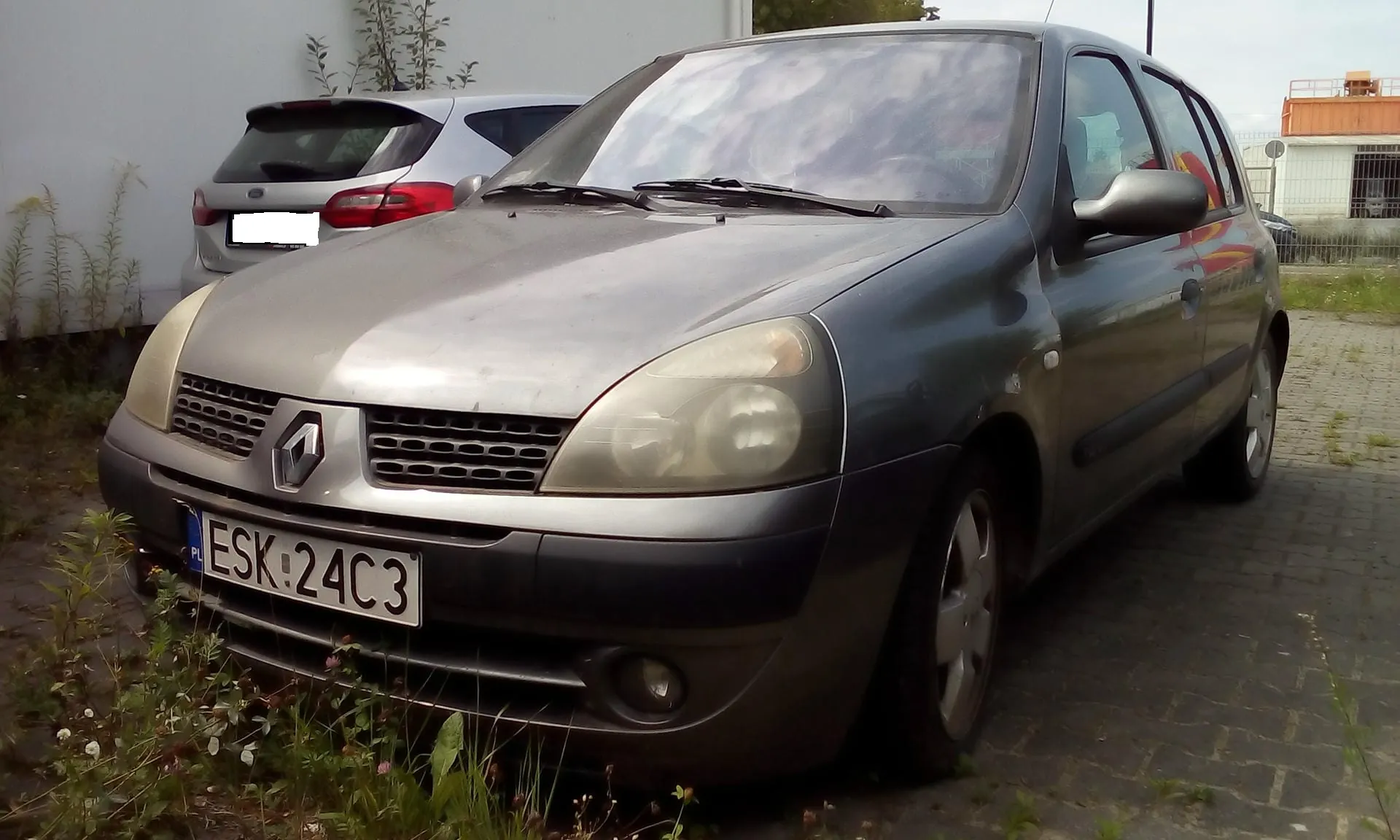 Srebrny samochód osobowy marki Renault zaparkowany na kostce brukowej. Widoczna jest przednia część pojazdu z tablicą rejestracyjną o numerze ESK 24C3. W tle znajduje się inny, jasny samochód oraz biała ściana budynku.