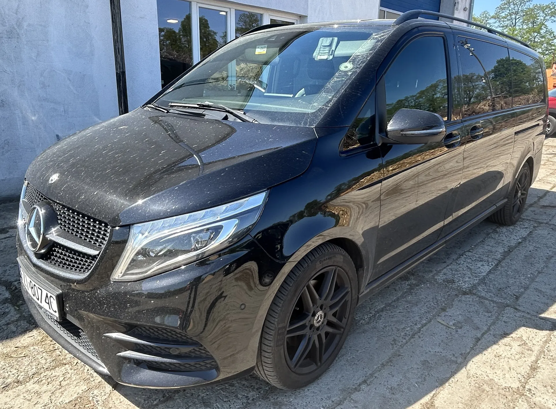 Czarny samochód osobowy typu van marki Mercedes-Benz zaparkowany na brukowanej powierzchni. Widoczna tablica rejestracyjna z numerem 1A 807 AC, chromowana atrapa chłodnicy z logo marki, czarne felgi, dachowy bagażnik oraz reflektory. W tle budynek z szarymi ścianami, dużymi oknami i niebieską bramą. Na szybie przedniej naklejki oraz odbicie drzew i nieba.