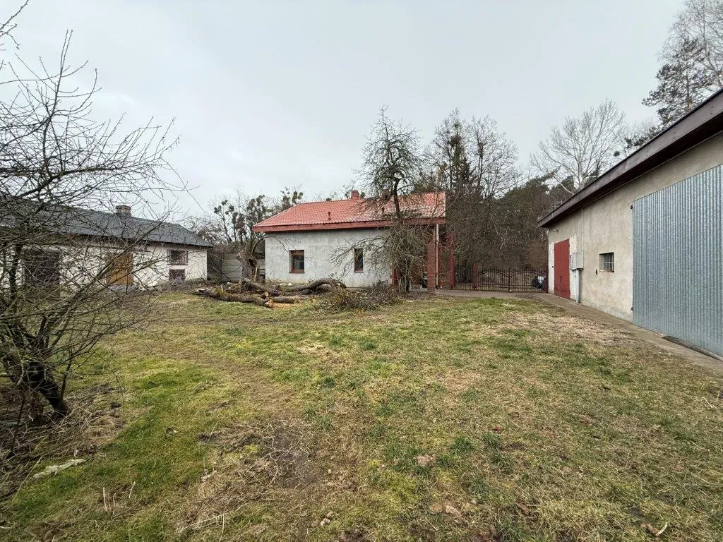 Zdjęcie przedstawiające podwórze z kilkoma budynkami gospodarczymi. W centrum znajduje się niewielki, biały dom z czerwonym dachem dwuspadowym. Po lewej stronie widoczny jest starszy, biały budynek z szarym dachem, a po prawej długa, biała hala z dużymi, szarymi metalowymi drzwiami. Na pierwszym planie znajduje się trawnik z kępami suchej trawy i ziemi, a po lewej stronie bezlistne drzewo. W tle widać las i zachmurzone niebo.