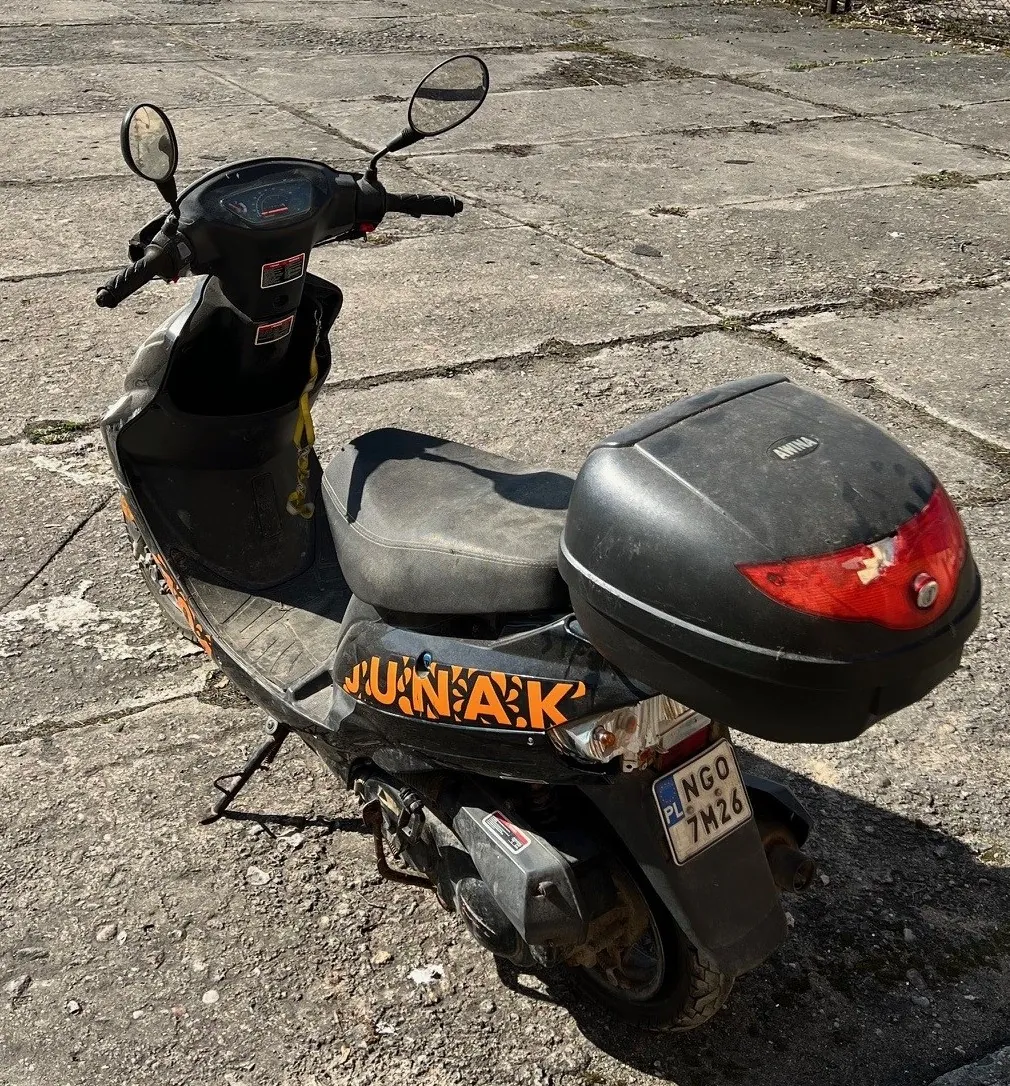 Czarny skuter zaparkowany na betonowej powierzchni. Skuter posiada pomarańczowy napis "JUNAK" na boku, szare siedzenie, czarny bagażnik z czerwonym światłem tylnym oraz tablicę rejestracyjną PL NGO 7H26. Widoczne są lusterka boczne, kierownica i stojak. Tło stanowi pęknięta betonowa nawierzchnia z drobnymi plamami mchu.