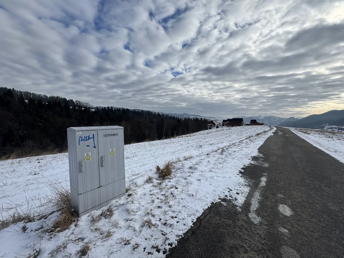 Krajobraz zimowy z asfaltową drogą prowadzącą w stronę kilku domów na wzgórzu. Po lewej stronie drogi stoi szara, metalowa szafka elektryczna z napisem '22796/RD' oraz żółtym trójkątnym znakiem ostrzegawczym przed wysokim napięciem. Teren wokół drogi jest pokryty cienką warstwą śniegu i suchą trawą. W tle widoczne są zalesione wzgórza pod zachmurzonym niebem.