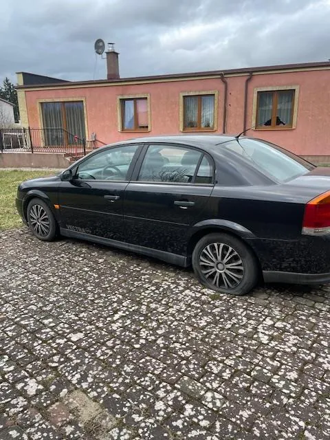 Czarny samochód osobowy typu sedan zaparkowany na brukowanej powierzchni. W tle różowy dom z kilkoma oknami, kominem i anteną satelitarną na dachu. Niebo jest zachmurzone.