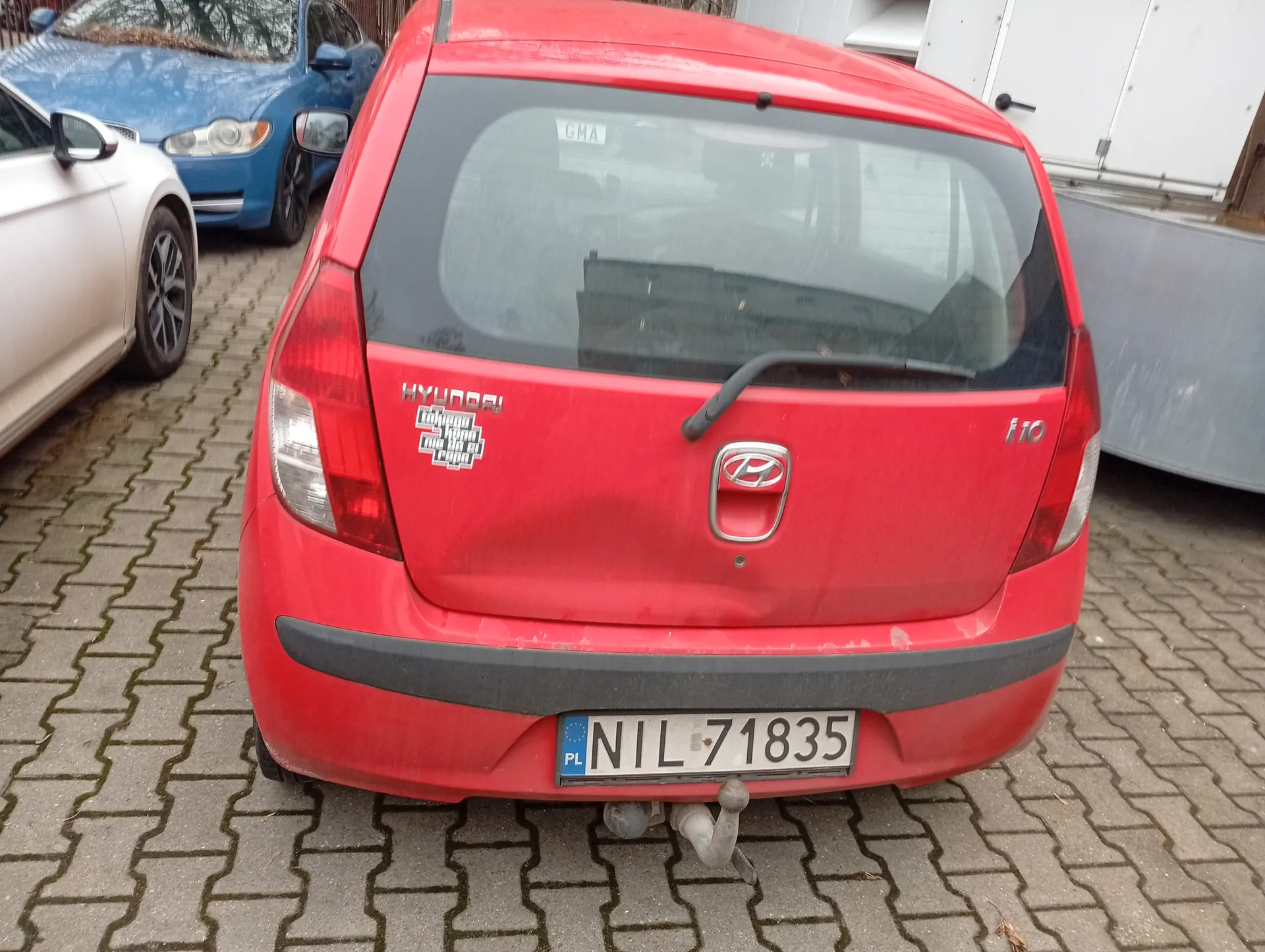 Czerwony samochód Hyundai i10 z tyłu, z tablicą rejestracyjną PL NIL 71835. Na bagażniku widoczny napis HYUNDAI, logo marki, oznaczenie i10 oraz naklejka z czarno-białym wzorem. W tle biały i niebieski samochód zaparkowane na brukowanej powierzchni. Po prawej stronie metalowa konstrukcja i biała ściana budynku. Podwozie czerwonego samochodu wyposażone w holownik.