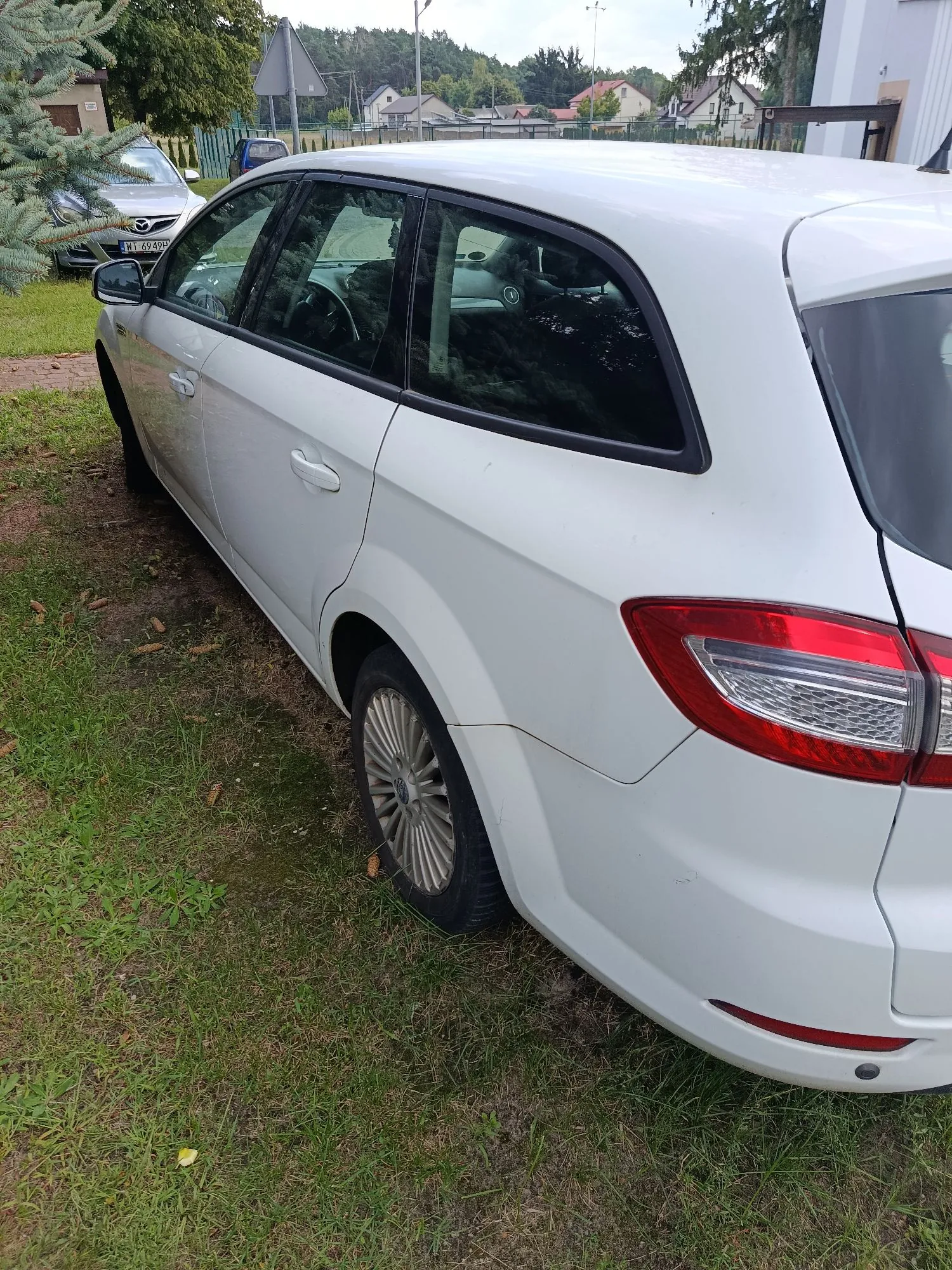 Biały samochód typu kombi zaparkowany na trawie. Widoczny jest prawy bok pojazdu, tylna lampa oraz fragmenty kół. W tle widać inne samochody, domy jednorodzinne oraz drzewa pod zachmurzonym niebem.