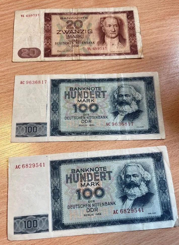 Trzy banknoty z Niemieckiej Republiki Demokratycznej (NRD) ułożone pionowo jeden nad drugim na drewnianym blacie. Górny banknot ma nominał 20 Mark (Zwanzig Mark) w kolorze brązowym z portretem mężczyzny. Dwa dolne banknoty mają nominał 100 Mark (Hundert Mark) w kolorze niebiesko-szarym z portretem Karola Marksa. Na każdym z banknotów widnieje napis 'BANKNOTE', 'DER DEUTSCHEN NOTENBANK DDR' oraz data 'Berlin 1964'. Widoczne są również numery seryjne: YL 659737 na banknocie 20 Mark, AC 9636817 na pierwszym banknocie 100 Mark oraz AC 6829541 na drugim banknocie 100 Mark.