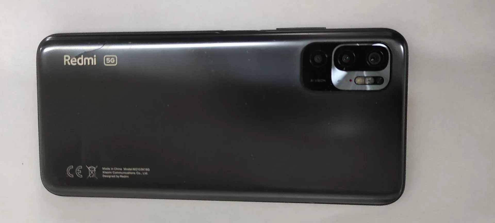 Tylna obudowa czarnego smartfona marki Redmi. W lewym górnym rogu znajduje się biały napis 'Redmi' oraz logo '5G'. W prawym górnym rogu umieszczono moduł z trzema obiektywami aparatów oraz lampą błyskową. W lewym dolnym rogu widoczne są oznaczenia certyfikacji CE oraz drobny tekst techniczny: 'Made in China Model M2103K1B0 Xiaomi Communications Co., Ltd. Designed by Redmi'.