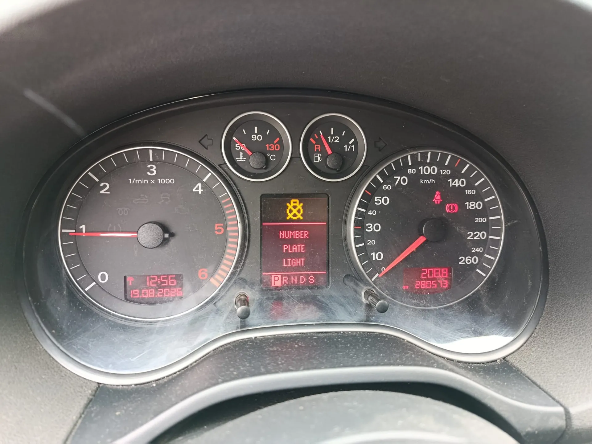 Zbliżenie na deskę rozdzielczą samochodu. Widoczny jest obrotomierz po lewej stronie, wskaźnik temperatury cieczy chłodzącej i poziomu paliwa na górze oraz prędkościomierz po prawej stronie. Na środkowym wyświetlaczu widnieje czerwony napis: 'NUMBER PLATE LIGHT' oraz żółta ikona ostrzegawcza. Na dole wyświetlacza obrotomierza widoczna jest data '19.08.2021' i godzina '12:56', a na wyświetlaczu prędkościomierza przebieg '208828.0573'.