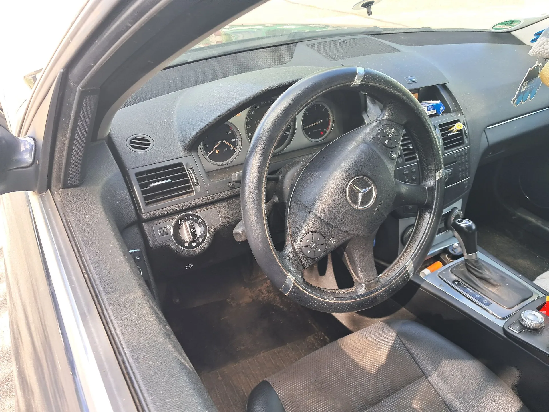 Wnętrze samochodu marki Mercedes-Benz widziane z perspektywy otwartych drzwi kierowcy. Widoczna jest czarna kierownica z logo marki, deska rozdzielcza z analogowymi wskaźnikami prędkości i obrotów, kratki nawiewów oraz konsola środkowa z dźwignią zmiany biegów. Fotel kierowcy jest wykonany z czarnej, perforowanej skóry.