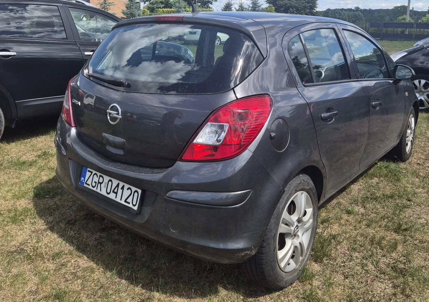 Szary samochód marki Opel Corsa zaparkowany na trawie. Widok z tyłu i z boku. Pojazd posiada polską tablicę rejestracyjną o numerze ZGR 04120.