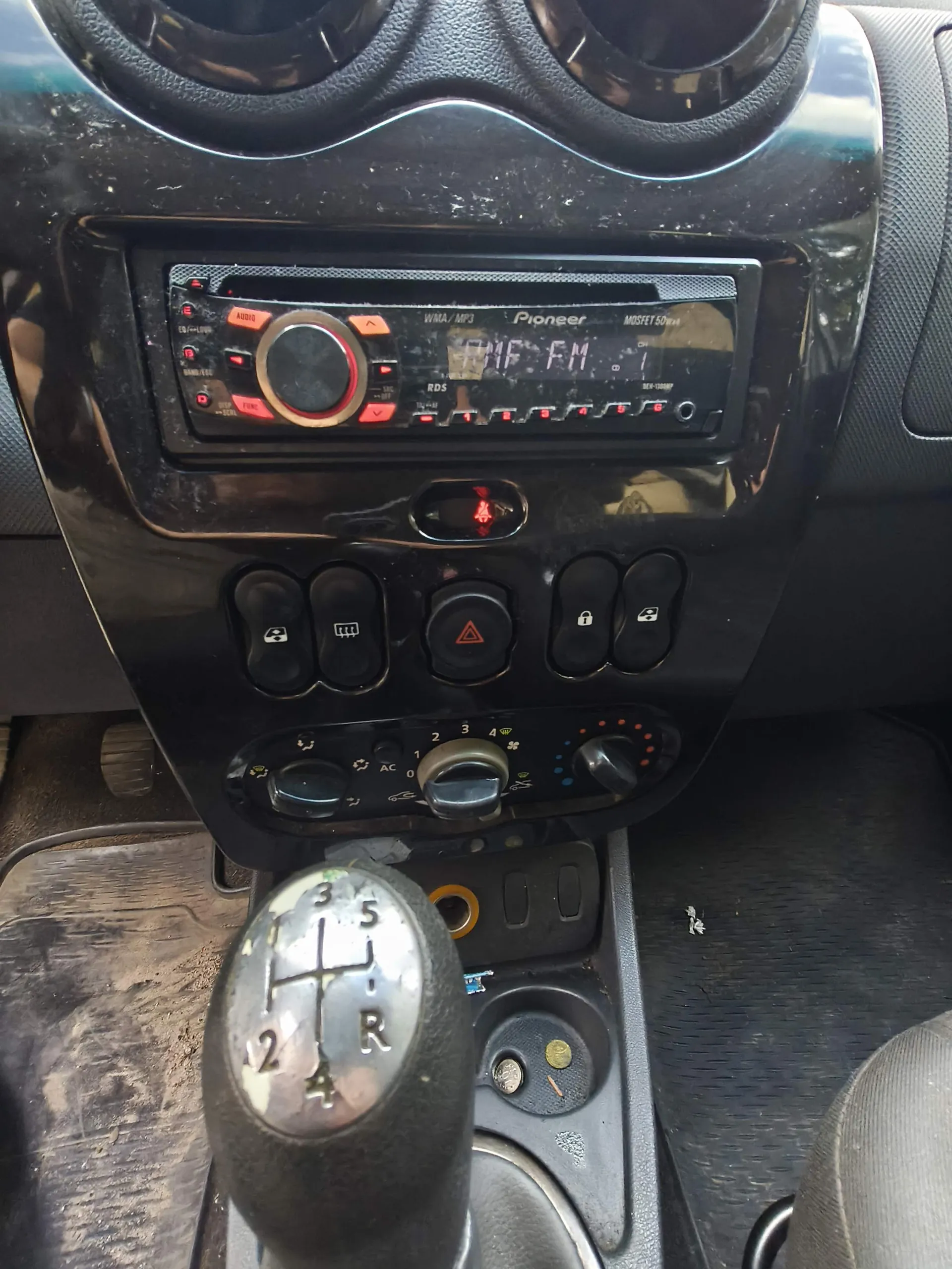 Wnętrze samochodu z widokiem na konsolę środkową. Na górze znajduje się radio marki Pioneer z wyświetlaczem LCD pokazującym napis 'FM'. Poniżej radia znajdują się przyciski sterujące, w tym czerwony przycisk świateł awaryjnych. Jeszcze niżej umieszczone są pokrętła klimatyzacji. Na pierwszym planie widoczna jest gałka lewarka zmiany biegów z oznaczeniami biegów 1-5 oraz wstecznego 'R'.